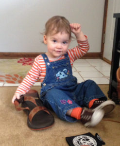Image shows a toddler sitting on the floor in denim overalls and a striped shirt signing "Daddy" while holding a large sandal.